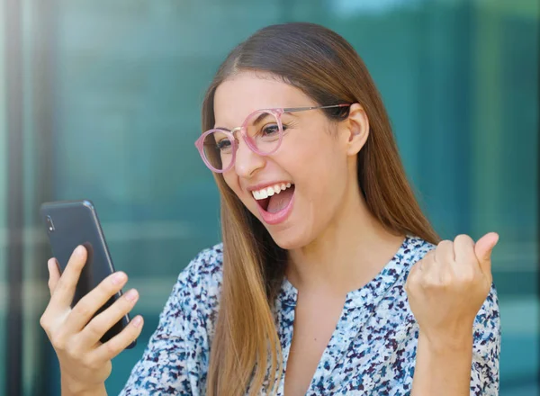 Mujer joven emocionada con gafas revisando su teléfono inteligente al aire libre —  Fotos de Stock