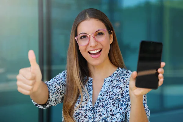 Emocionado joven mujer muestra su teléfono inteligente y pulgar hacia arriba al aire libre . —  Fotos de Stock