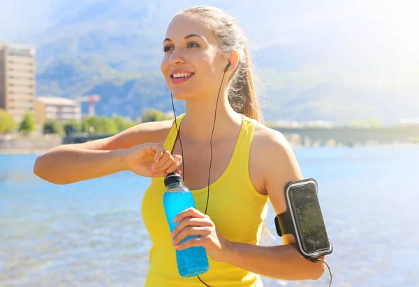 Feliz atleta femenina alegre con brazalete para teléfono inteligente botella de bebida de energía abierta después del entrenamiento diario y mirando al aire libre . — Foto de Stock