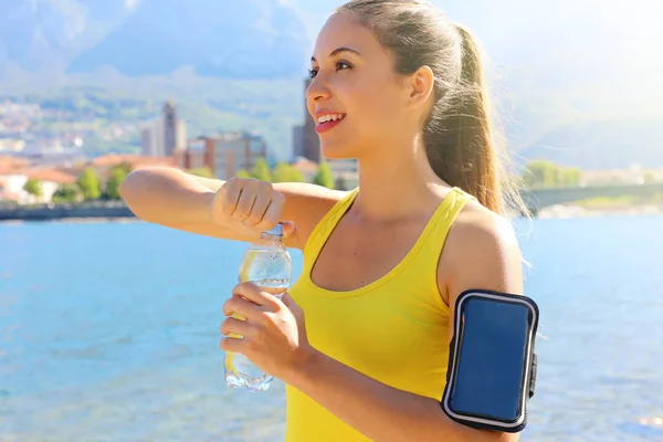 Thirsty fitness woman opens bottle of water after training outdoor. Fit woman using smartphone fitness app on armband as activity tracker.