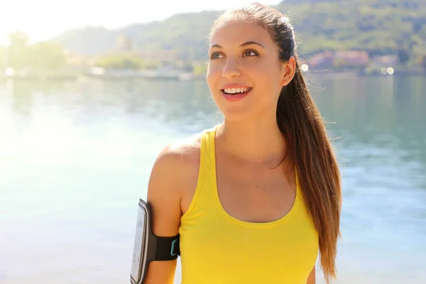 Atractiva mujer fitness con brazalete para teléfono inteligente mirando a un lado el espacio de copia con luz del sol de la mañana y el lago en el fondo . — Foto de Stock