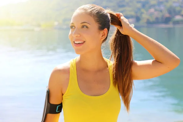 Retrato de una chica fitness sonriente con banda de brazo fijando su cabello mientras está de pie al aire libre mirando al espacio de copia lateral . —  Fotos de Stock