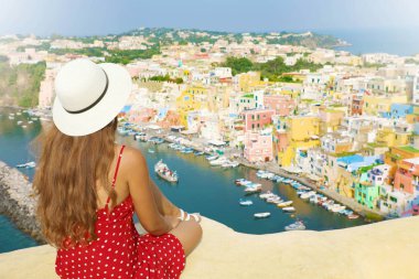 Beautiful young female model with hat in Procida Island with Marina di Corricella harbor and the village on the background, Procida, Italy. clipart