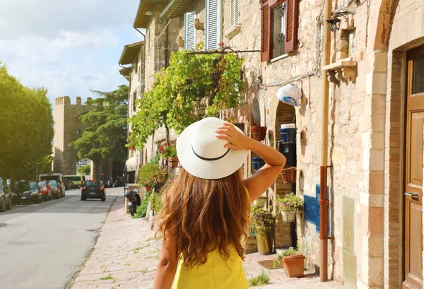 Bella giovane donna con abito giallo e cappello passeggiando nella tipica via Assisi, Italia. Vista posteriore di felice ragazza allegra in visita in Italia centrale . — Foto Stock