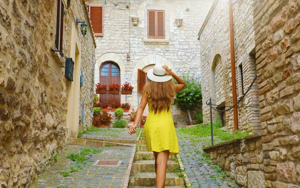 Krásná zvědavá mladá žena se žlutými šaty a kloboukem jde nahoru po ulici v Assisi v Itálii. Zadní pohled na šťastnou veselou dívku na návštěvě střední Itálie. — Stock fotografie