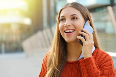 Oldukça neşeli genç bir bayan telefonda konuşuyor ve dümdüz ileriye bakıyor. Akıllı telefonu kulağına yakın tutan kız dışarıda tek başına duruyor. Arka planda modern şehir manzaralı gün ışığı.