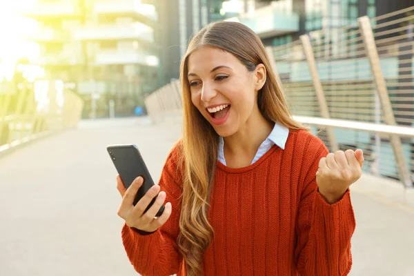 Emocionada joven mirando su teléfono en la calle. Chica ganadora celebrando con móvil en su mano afuera . —  Fotos de Stock