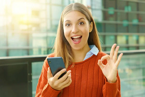 Feliz hermosa mujer de otoño sosteniendo el teléfono inteligente haciendo Ok signo en la cámara. Emocionado joven satisfecho utilizando el teléfono inteligente al aire libre . —  Fotos de Stock