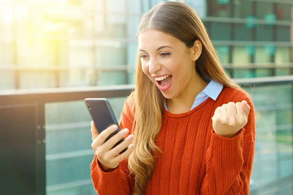 Feliz mujer emocionada usando un suéter rojo leyendo buenas noticias en línea afuera en la calle en otoño . —  Fotos de Stock