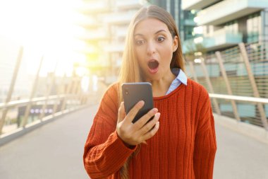Şok olmuş ve şaşırmış genç bir kadın modern Avrupa şehrinde akıllı telefonuyla mesajlaşıyor ve konuşuyor..