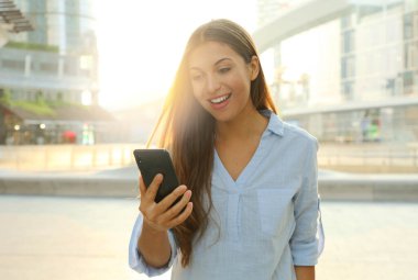 Mutlu neşeli kız modern şehirde sabah sabah akıllı telefonunu kontrol ediyor..