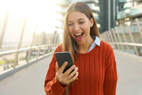 Mujer eufórica mirando su teléfono inteligente con paisaje urbano moderno en el fondo . —  Fotos de Stock
