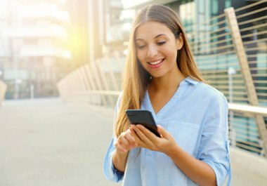 Modern şehir caddesinde dururken akıllı telefonunda yazan mutlu iş kadını. Boşluğu kopyala.