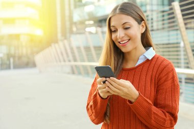 Modern şehir caddesinde dururken akıllı bir telefonla mesaj yazan ya da okuyan mutlu bir öğrenci kadın. Boşluğu kopyala.