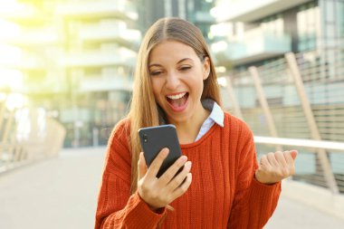 Açık havada akıllı telefonunu izleyen heyecanlı bir kadın. Neşeli kadın modern şehirde akıllı telefonunu kontrol ediyor..