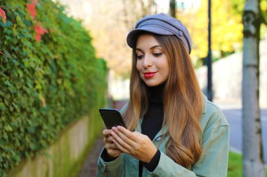 Şapkalı ve ceketli güzel genç bir kadın açık havada cep telefonuyla mesajlaşıyor..