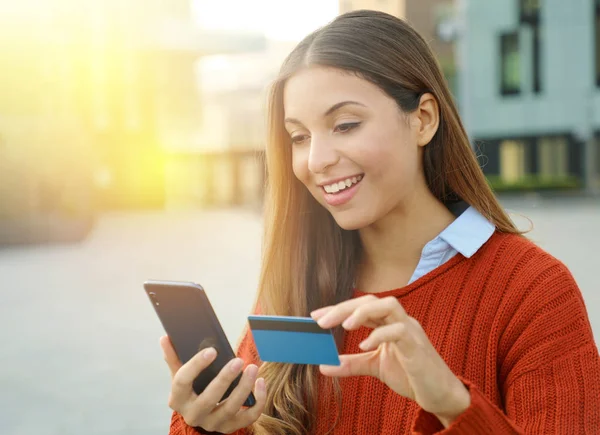 Retrato de uma mulher bonita segurando um cartão de crédito para comprar on-line com um telefone inteligente ao ar livre . — Fotografia de Stock