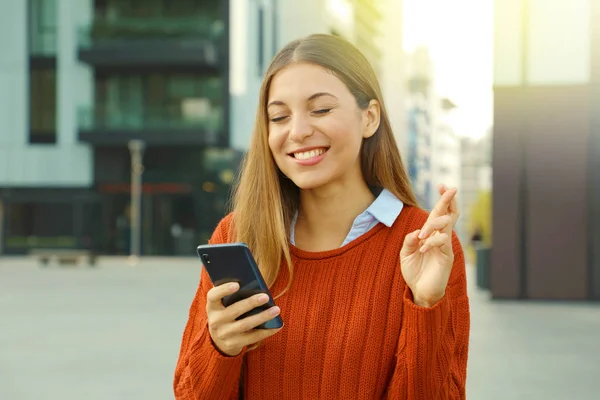 Chica bastante esperanzada cruzando los dedos y muerde el labio sosteniendo un teléfono inteligente esperando buenas noticias al aire libre . —  Fotos de Stock