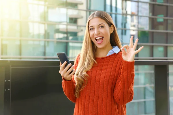 Feliz hermosa mujer de otoño sosteniendo el teléfono inteligente haciendo Ok signo en la cámara. Emocionado joven satisfecho utilizando el teléfono inteligente en la ciudad moderna . —  Fotos de Stock