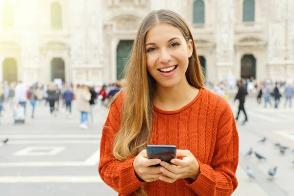 Mujer joven feliz con suéter en las buenas noticias en su teléfono móvil mirando a la cámara en la plaza de la ciudad . —  Fotos de Stock