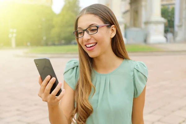 Sonriendo joven mujer de negocios con gafas haciendo videollamada con su teléfono inteligente al aire libre . — Foto de Stock
