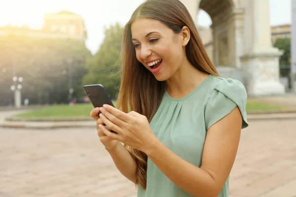 Sorprendida eufórica mujer alegre viendo su teléfono inteligente en la plaza de la ciudad . —  Fotos de Stock