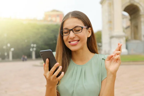 Chica bastante esperanzada con gafas cruzando los dedos y sosteniendo un teléfono inteligente esperando buenas noticias al aire libre. Mujer joven con los dedos cruzados y teléfono inteligente que desea lo mejor fuera . —  Fotos de Stock