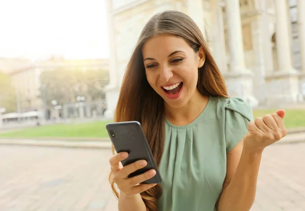 Mujer alegre eufórica comprobando su teléfono inteligente en la plaza de la ciudad . —  Fotos de Stock