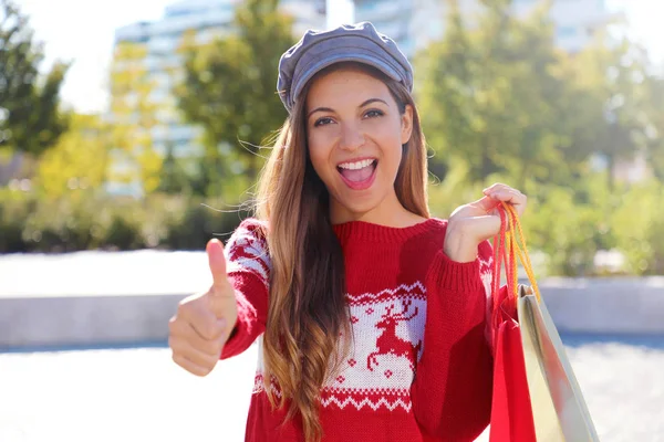 Happy excited christmas woman holding shopping bags gives thumb up outdoors.