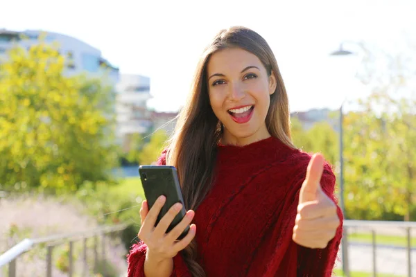 Cheerful laughing young woman with mobile phone giving thumb up outside.
