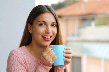 Kameraya bakan mutlu genç bir kadın elinde sıcak bir kahve tutuyor..
