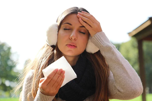 Mujer joven con orejeras y bufanda sufriendo migraña dolor de cabeza sosteniendo tejido al aire libre . — Foto de Stock