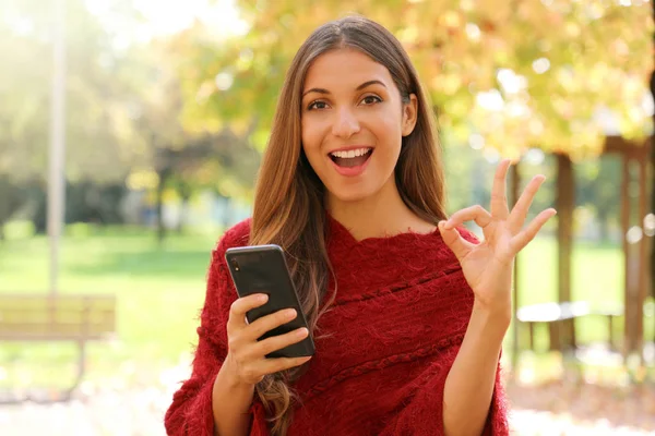 Hermosa mujer joven sosteniendo el teléfono inteligente y haciendo signo OK al aire libre en otoño o invierno temporada . —  Fotos de Stock
