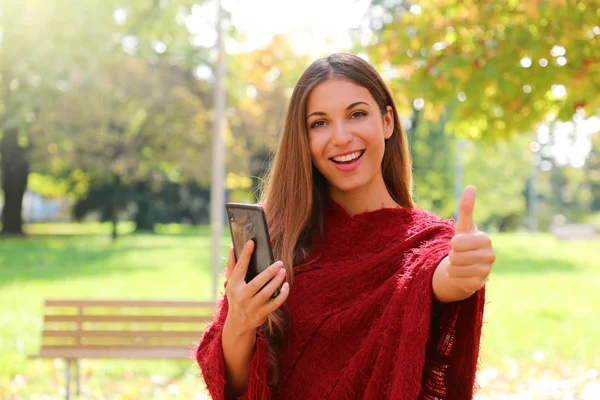 Retrato de una mujer de moda usando un teléfono inteligente mostrando el pulgar hacia arriba en el parque de la ciudad —  Fotos de Stock