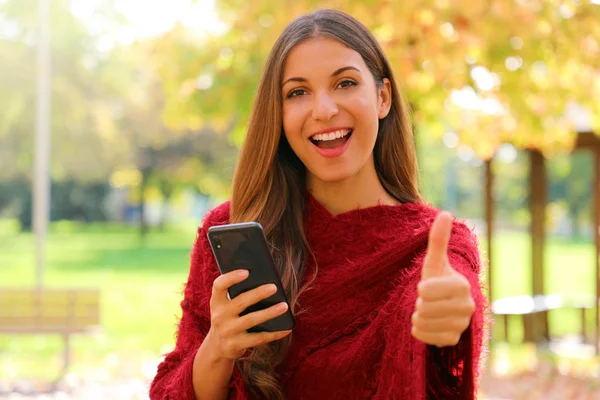 Atractiva mujer de la moda usando el teléfono inteligente muestra el pulgar hacia arriba al aire libre en otoño . —  Fotos de Stock