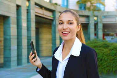 Dışarıda cep telefonu kullanan başarılı genç ve mutlu bir iş kadını. Ofis kıyafetli güzel bir kadın elinde akıllı telefon, kameraya gülümsüyor. Taşınabilir internet kavramı.