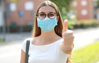 Koruyucu maske takan iyimser bir kızın portresi başparmağını şehir sokaklarında gösteriyor.
