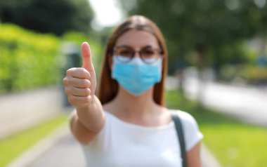 Young student girl with surgical mask showing a sign of like outdoor. Focus on the hand. clipart