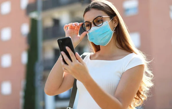 City lifestyle stylish hipster girl with surgical mask reading a message on mobile phone in street