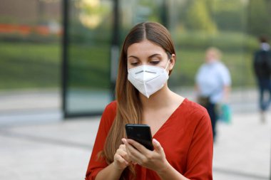 KN95 FFP2 maskeli genç kadın Coronavirus Hastalığı 2019 'a tepki olarak Şehir Caddesinde Cep Telefonu Uygulaması ile Kişinin İzlenmesine ve Kişiliğine Yardım Ediyor
