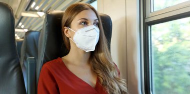 Travel safely on public transport. Young woman with KN95 FFP2 face mask looking through train window. Train passenger with protective mask travels sitting in business class looking through the window.