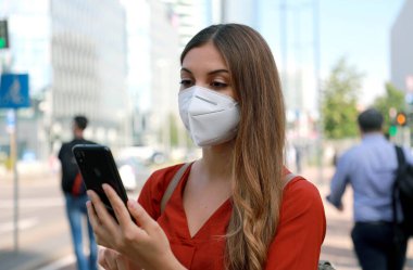 KN95 FFP2 maskeli bir iş kadını modern şehir caddesinde arka planda insanların olduğu bir akıllı telefonla yürüyor.