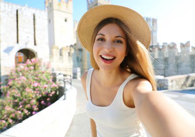 Gülümseyen genç turist kadın Sirmione, İtalya 'da kendi portresini çekiyor. Güzel moda kızı arka planda Sirmione Kalesi ile resim çekiyor, Garda Gölü, İtalya.