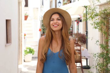 Eski kasabadaki antik caddede neşeli turist kadın portresi. Hasır şapkalı ve mavi elbiseli çekici bir kadın Avrupa 'da tek başına tatilin tadını çıkarıyor. Turizm ve seyahat kavramı.