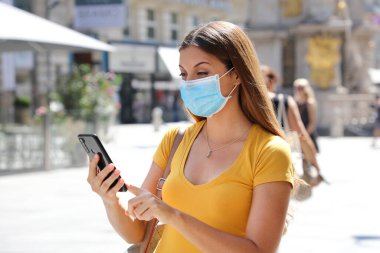 COVID-19 koruyucu maskeli iyimser genç kadın cep telefonundan iyi haberler okuyor.