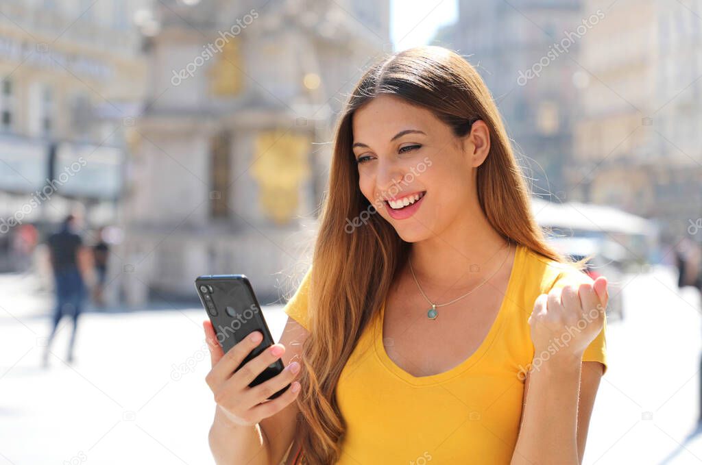 Happy excited satisfied woman holding smart phone reading good news outdoor and celebrating in city street