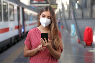 KN95 FFP2 maskeli genç bir kadın istasyonda bekliyor. Beyaz kadın tren istasyonunda akıllı telefon kullanıyor..