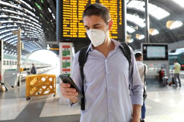 Tren istasyonunda KN95 FFP2 maskesi takan genç bir iş adamı. Tren istasyonunda akıllı telefon kullanarak gelen gidiş saatlerinin gerisinde kalan beyaz bir adam..