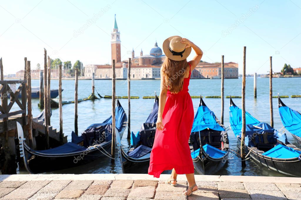 Back view of beautiful girl in red dress walking in Venice with gondolas moored