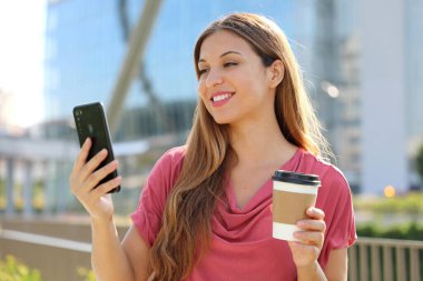 Gülümseyen Brezilyalı bir kadın pembe bluz giyip açık hava telefonuyla arıyor, elinde kahve fincanı tutuyor.